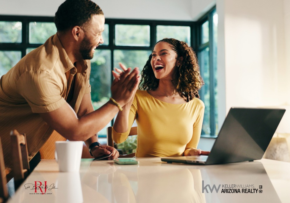 Two coworkers share a high-five across a bright desk, with a laptop in a modern office setting, celebrating a win in real estate.