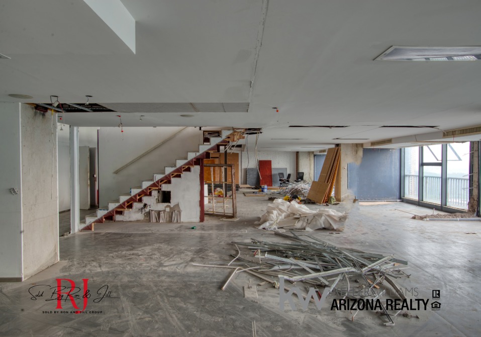 Open-plan interior under renovation with a wooden staircase on the left, construction debris on the floor, and large windows along the right wall.