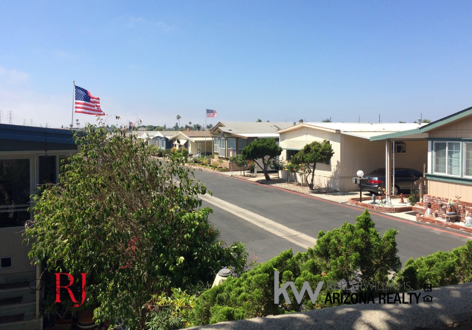 Wide view of a manufactured home community along a paved street, with American flags on flagpoles, shrubs in the foreground, and blue sky above.