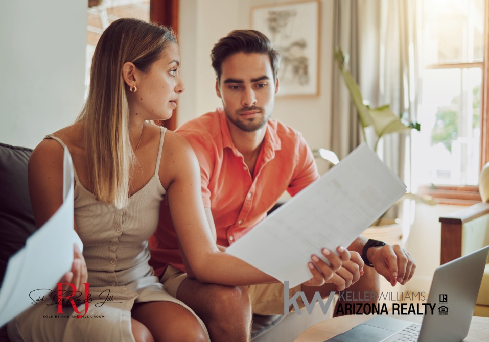 Stressed 001 - Sold By Ron And Jill Group - Sold By Ron And Jill Group Couple sitting on a couch examining a large property document during a real estate consultation.